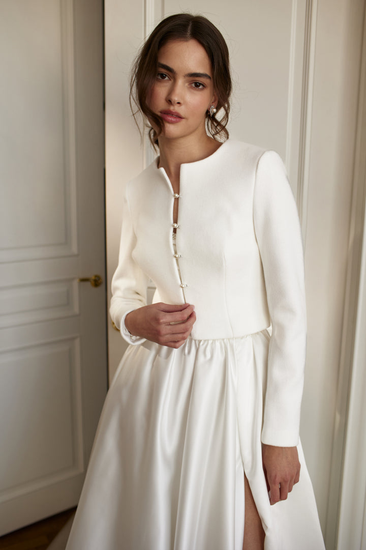 A bride wearing an ivory wedding jacket with pearl buttons and a white maxi dress, standing indoors