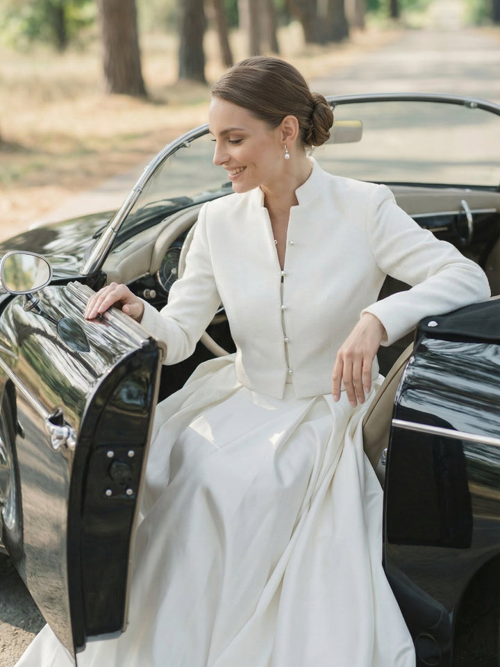Bride wearing a cropped white wedding jacket while sitting beside a vintage car