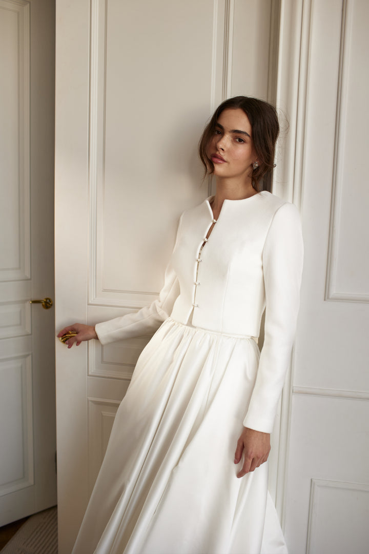 A bride wearing a white wedding coat over a long flowing gown, standing by a doorway