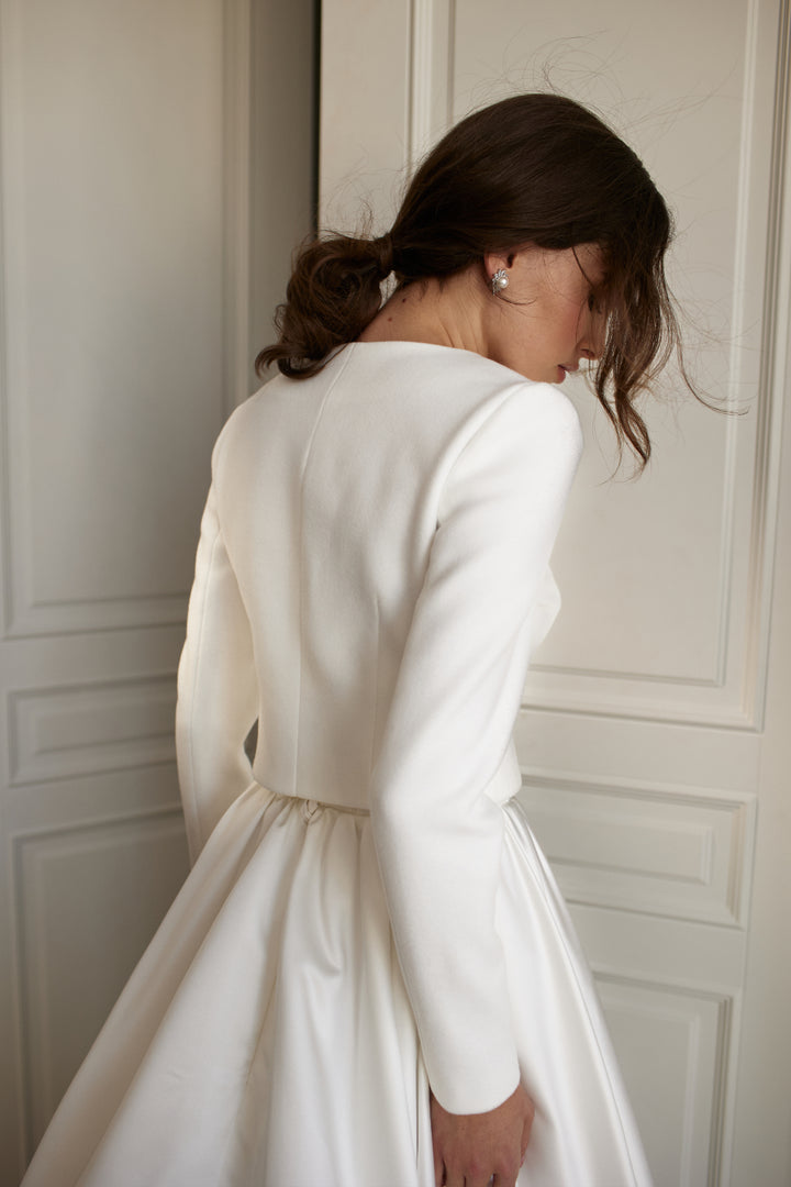 Back view of a bride wearing an ivory wedding jacket over a white wedding dress indoors
